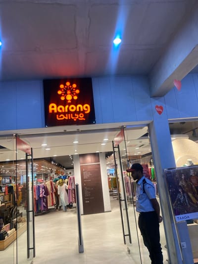 An image depicts the entrance to an Aarong retail store, identified by its illuminated orange sign featuring the brand name in English and Bengali, along with a circular, molecular-like logo. This store is located in Narayanganj, Bangladesh.

The entrance consists of glass double doors with black metal frames, flanked by grey security sensor pillars. A male security guard stands to the right of the entrance, facing inwards, wearing a light blue short-sleeved uniform shirt with the number "03" on the right sleeve, dark trousers, and a dark baseball cap.

Inside the brightly lit store, multiple customers are visible, primarily women, browsing racks of colorful traditional clothing items such as saris and kurtis. On the left side of the entrance, display shelves hold various accessories and home goods. A prominent vertical brown sign within the store lists sections including "TAAGA & MAN," "TAAGA WOMAN," "EARTH," "AARONG," and "SIXTH." To the right inside the store, a large, textured white circular display is present. Overhead, the ceiling is light grey concrete with visible marks and two bright blue-toned lights. Red heart-shaped decorations are affixed to a pillar and wall to the right of the entrance, one bearing the text "I Love Aarong." A "TAAGA" brand poster is positioned to the immediate right of the entrance, partially obscuring a power outlet. The store floor is composed of light-colored tiles.