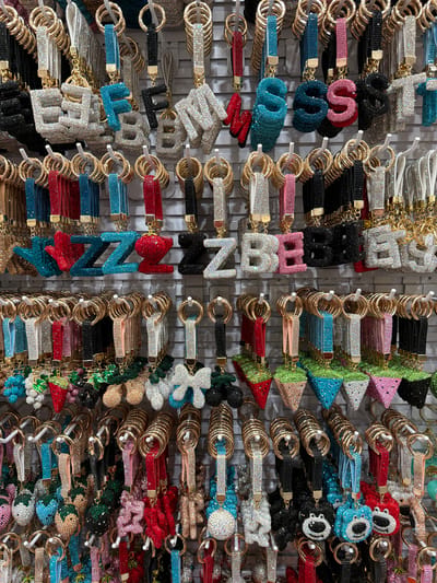 This image shows a wall display of numerous bejeweled keychains. The keychains are arranged in rows and columns, with many featuring letter shapes, geometric patterns, or abstract designs. They are made of small, reflective stones, giving them a sparkly appearance. Colors include silver, gold, black, pink, blue, red, and green. Some keychains have a tassel-like element at the bottom. The background appears to be a white, gridded display wall, typical of a retail environment. The lighting is bright and even, suggesting an indoor setting like a shop. The sheer quantity and variety of the keychains indicate they are for sale. No people are visible. The scene is static and offers no information about the time of day or weather. The location context suggests this display is in Beyoğlu, Turkey, likely in a store selling accessories. No visible text is present on the keychains or the display.