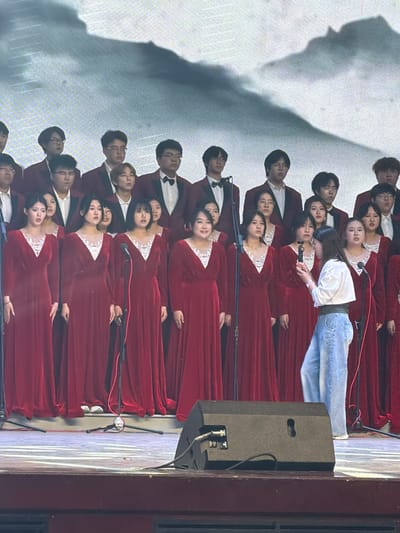 This image depicts an indoor choral performance or rehearsal in Guta, China. A large group of choir members, consisting of both men and women, is arranged in multiple rows on a stage. Most performers are uniformly dressed in long, deep red gowns with intricate white lace or embroidered necklines. Male choir members wear red jackets over white shirts with black bow ties. Their expressions are mostly focused and serious, typical of performers.
In the center of the front rows, a woman with long dark hair, wearing a white long-sleeved top and light blue jeans, stands and appears to be interacting with a microphone on a stand. She might be a conductor, a soloist, or a member of the event staff. Numerous microphones on stands are positioned throughout the choir rows.
To the right side of the frame, a large, dark speaker or monitor is visible, along with sections of red stage curtains or drapes. The background features a large, light-colored screen or wall onto which a prominent, abstract, dark shadow, resembling clouds or a natural formation, is projected. Red and black audio cables are visible on the stage floor. The setting suggests a formal event in an auditorium or performance hall, under artificial lighting, indicating it is likely an evening event.