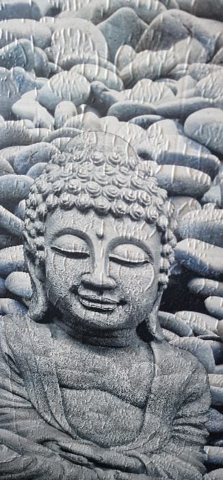 The image depicts a close-up view of a grey-toned, relief-style representation of a Buddha figure. This image does not show a location in the city of Blaye, France; it appears to be a photograph of a surface such as a tile, wallpaper, or decorative panel.
The central subject is the head and upper torso of a Buddha. The Buddha's face is rendered with closed eyes and a slight, serene smile. The head is covered with tight, small curls, characteristic of Buddha depictions. The visible torso is draped in robes, indicated by sculpted folds. The entire Buddha figure has a rough, textured appearance, suggesting stone or concrete.
The background consists of numerous rounded, grey stones or pebbles of various sizes, creating a textured field around the Buddha. The stones also exhibit a somewhat rough surface texture, consistent with the Buddha figure. Both the Buddha and the surrounding stones are monochromatic, primarily in shades of grey, with subtle variations in tone providing depth and dimension. There are reflective highlights across the textured surfaces, suggesting a glossy or semi-glossy finish on the material being photographed. No living individuals, active movements, or direct interactions are visible.