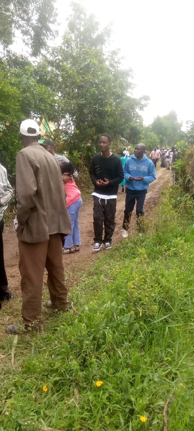 The image shows a group of people standing on a dirt path in a rural area, likely in a village or small town. The location, based on the provided coordinates, is near the Burundu market in Embu County, Kenya. The people are all African in appearance, and they are dressed in casual clothing. There are two men standing in the foreground, one wearing a brown jacket and white cap, and the other wearing a blue shirt and black pants. Behind them, three other individuals can be seen: one young man in a black sweater and black pants, a young woman in a pink shirt and blue jeans, and another man in a light blue shirt. The environment is characterized by a dirt path lined with tall green vegetation and trees on either side. The path is likely leading to or from a market or community gathering place, and the people are likely returning home or heading to a nearby location. The overall mood appears casual and peaceful.
