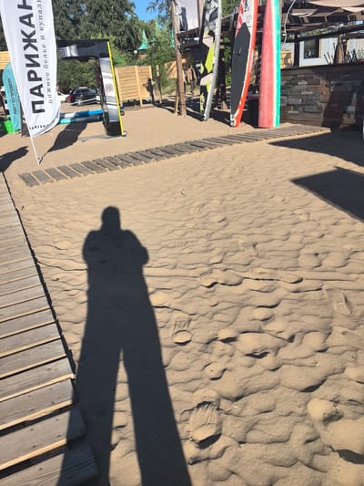 A person's shadow is cast across a sandy beach area, suggesting a sunny day. In the background, several paddleboards are propped up against a wooden structure, indicating a recreational water sports setting. A large white banner with Cyrillic text hangs prominently on the left, reading "ПАРИЖАН" (Parizhanka) and below it, "нижнее белье и купальники" (lingerie and swimwear). To the left of the banner, another smaller banner reads "АВТОЛИДЕР" (Autolider). A black SUV is parked in the mid-ground, and wooden planks create a pathway through the sand. The overall scene is likely a beachside establishment or rental point for water sports gear, possibly in Tolyatti, Russia.
