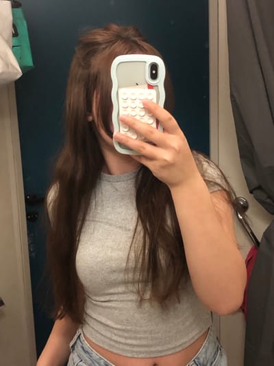 An individual, likely female, is depicted in a mirror selfie within an indoor setting. The person has long, wavy brown hair, partially styled in a half-up bun, and is wearing a light grey short-sleeved crop top paired with light-wash blue denim jeans. Their face is obscured by the smartphone they are holding, which is an iPhone X, XS, or 11 Pro series device, housed in a pale blue or mint green wavy-edged case. The back of the phone case features a grid of approximately 18-20 small, circular white button-like protrusions. A red accent is visible near the phone's camera module. The background, reflected in the mirror, shows a dark blue/teal door with a black handle on the left. To the right, a grey curtain or fabric is visible alongside a light-colored wall featuring a silver-colored metal hook or bar from which a small red fabric item is partially suspended. The specific location within the city of Saverne, France, cannot be determined from the visual evidence in the image.