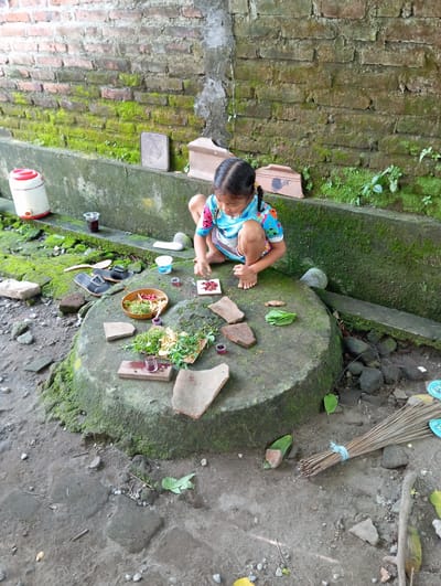 A child is seated on a large, moss-covered stone structure in Baron, Indonesia. The surrounding area is a mix of brick and concrete, also covered in moss. The child is surrounded by various objects including a wooden bowl with plants, leaves, small cups, and fragments of broken pottery. A broom rests in the background.