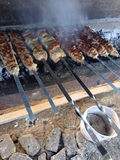 The image captures a close-up, elevated view of eight metal skewers of meat grilling over hot charcoal, generating smoke, in Kasri Arifan, Uzbekistan. The skewers hold two types of meat: elongated ground meat preparations and cubed pieces of meat and fat. The meat is visibly cooking and browning from the heat of the glowing, ash-covered coals. The grill structure appears to be a simple, dark-toned charcoal brazier, possibly constructed from brick or concrete. Below the grill, the ground surface consists of loose soil and scattered fragments of concrete or bricks. A white, cylindrical bucket, possibly containing ash or debris, is partially visible in the lower right foreground. No individuals are present in the scene.