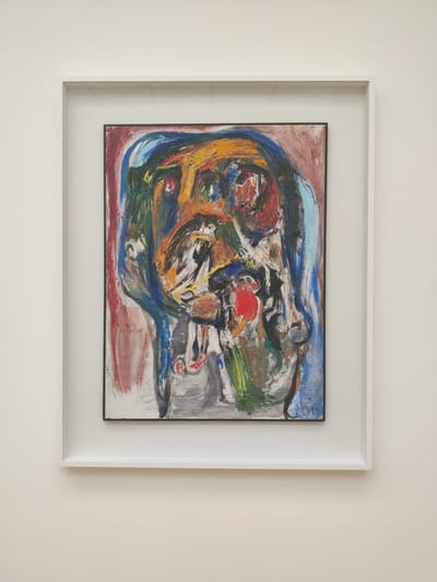An abstract expressionist painting is framed in a white shadow box frame and mounted on a white wall. The painting is characterized by bold, energetic brushstrokes and a vibrant color palette. The primary colors used are red, yellow, blue, green, black, and white, applied in thick impasto. The composition is dynamic and chaotic, with swirling lines and forms that suggest movement and emotion.
The subject matter is abstract, with no clear representational figures or objects. However, the arrangement of colors and shapes evokes a sense of a tumultuous scene, perhaps a landscape in motion or an internal emotional state. There are areas that could be interpreted as stylized human forms or faces, particularly with the use of red and black pigments. A prominent red circular shape near the center might suggest a focal point or a dynamic element within the composition. The background appears to be a mix of blues and grays, with hints of red and brown at the edges.
There is some visible text in the upper right corner of the painting, appearing to be a signature or inscription in blue paint. The text is difficult to decipher fully due to the abstract nature of the painting and the quality of the image, but it seems to consist of a few letters.
The overall impression is one of intense energy and emotion, typical of abstract expressionism. The painting is displayed in a modern interior, suggested by the clean white wall and the minimalist shadow box frame. The lighting is neutral, indicating an indoor setting, possibly during the day. No specific weather conditions or time of day are discernible from the artwork itself. The location context of The Hague, Netherlands, does not directly inform the visual content of the abstract painting.