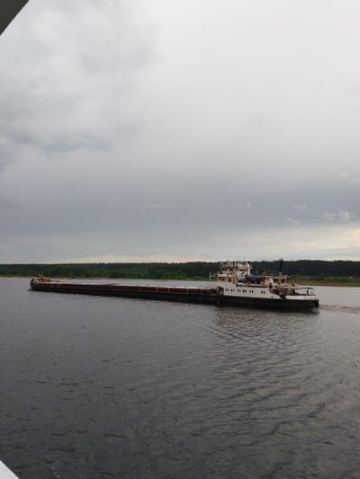 An overcast sky hangs over a wide body of water, likely the Volga River, in Myshkin, Russia. A long, dark barge is being pushed by a tugboat with a white superstructure. The tugboat is positioned at the rear of the barge. Dense green trees line the far bank of the river.