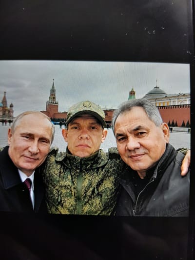 This media file shows a selfie of three men in front of the Kremlin. The man in the foreground on the right is Vladimir Putin, wearing a dark suit and a burgundy tie. To his left and slightly behind him is a man in a camouflage military uniform and a cap. Behind him, and to the left, is another man with gray hair, wearing a black jacket. The Kremlin, with its iconic red walls and towers, is visible in the background. The sky appears overcast, suggesting a cool or cloudy day. The overall scene conveys a sense of camaraderie or official gathering. There is no visible text in the image. The photo appears to have been taken with a digital camera or phone, and there are some minor artifacts from the screen display.