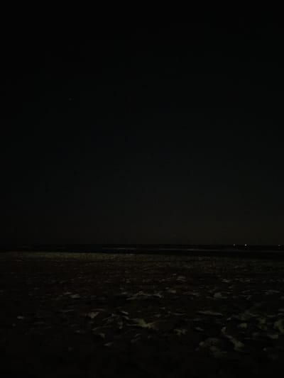 The image depicts a dark, expansive scene, likely taken at night. The foreground is dominated by what appears to be a sandy or uneven surface, with patches of lighter material scattered across it, possibly snow or ice. This textured area occupies the majority of the lower half of the frame.
Above the textured foreground, a dark horizon line separates it from a vast expanse of blackness that fills the upper portion of the image. This upper area is largely featureless, suggesting a clear night sky. There are a few very small, faint points of light visible in the upper portion of the frame, one slightly larger and brighter in the upper left quadrant, and two smaller, dimmer lights near the horizon on the right. These could be stars, distant lights, or possibly aircraft.
The overall impression is one of darkness and solitude. The lack of distinct features and the deep shadows make it difficult to ascertain the exact nature of the environment or the presence of any specific objects beyond the surface texture in the foreground. The scene is devoid of any visible human presence or activity. The location is identified as Unknown, Greece, but there are no visual cues within the image to support or refute this.