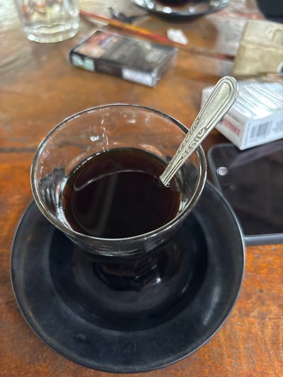 A close-up, high-angle shot shows a glass of dark coffee with a spoon resting inside, placed on a black saucer. The coffee is on a rustic wooden table. To the left of the coffee cup, a clear glass is visible, and further back, a pack of cigarettes lies on the table. To the right of the coffee cup, a white cigarette pack is present, along with a dark smartphone. The scene suggests a casual setting, possibly a cafe or a personal space, in Seuneubok Teungoh, Indonesia.
