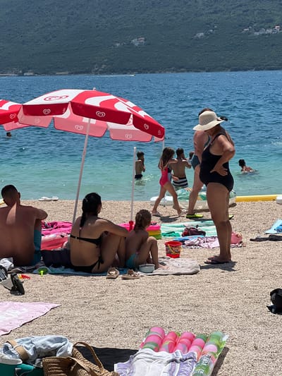 A sunny, outdoor scene takes place on a pebble beach in Baošići, Montenegro, with the turquoise waters of the Adriatic Sea in the background. In the foreground, a man and two women are seated on towels, facing away from the camera, while a young girl sits in front of them. A large, red and white umbrella shades the group. Children play in the shallow water, and a woman in a wide-brimmed hat stands near the water's edge, looking out at the sea. Several beach towels, bags, and inflatable toys are scattered on the sand. The mountainous coastline of Montenegro forms a lush green backdrop to the scene, with a few scattered houses visible on the slopes. The water is calm, with a gentle ripple, and the sky is clear and blue.