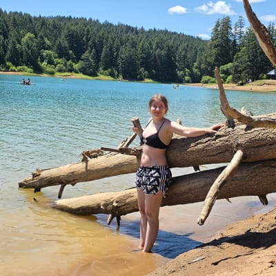 In Cherry Grove, Oregon, a young woman stands at the edge of a body of water, partially submerged, with her feet in the shallows. She is leaning against a large, fallen tree trunk that lies horizontally across the water and the shore. The woman is wearing a black bikini top and black and white patterned shorts. Behind her, the lake stretches out, bordered by a dense forest of evergreen trees that climb a hillside. The sky is blue with scattered white clouds. In the distance on the water, several people are visible, some on paddleboards and others in small boats. The shore is sandy with some exposed tree roots and debris. The overall scene depicts a sunny day at a natural lake with recreational activities.