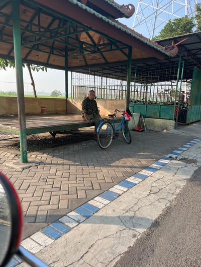 An elderly man sits on a wooden platform under a shaded pavilion with a green metal frame and a brown roof, adorned with ornate carvings. In front of the pavilion, a blue bicycle is parked on a paved walkway made of interlocking bricks. The walkway has a decorative border of blue and white tiles. Behind the pavilion, a green metal fence encloses a rack of bicycles. The background shows trees, a distant structure, and a bright sky. The scene appears to be in a public area in Karangtanjung, Indonesia, possibly a resting spot or waiting area.