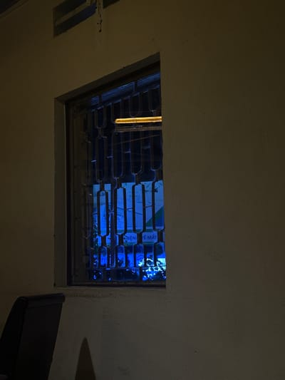 This media file shows a rectangular opening in a ceiling, possibly a skylight or ventilation shaft, covered by a metal grate. Through the grate, a bright blue light illuminates the interior. This blue light reflects off the grate and the ceiling, creating a dramatic and somewhat eerie effect.
The scene appears to be indoors, given the ceiling structure and the artificial lighting. The time of day is likely night, as the blue light is the dominant light source and contrasts sharply with the dim surroundings. The setting hints at a functional space, perhaps an industrial building or a storage area, given the visible grate and the somewhat rough texture of the ceiling.
Within the illuminated area, some objects are visible, though indistinct due to the lighting and distance. There are hints of shelves or containers, and possibly some foliage, suggesting it might be a storage area for plants or other items.
The most prominent visible text appears in the illuminated section, on what seems to be labels or signs. The text is in Vietnamese and includes "BÊN VỆ SINH" and "RẺ MẶT". These phrases suggest a context of hygiene or possibly a retail environment.
There is a shadow cast on the right side of the image, indicating a light source or object outside the frame. A dark, angular object, possibly a piece of furniture or equipment, is partially visible in the bottom right corner. The overall mood is mysterious and somewhat industrial, with the intense blue light being a key element. The location context of Thủ Đức, Vietnam, suggests that the Vietnamese text is consistent with the locale.