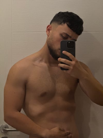 The image is a medium shot of a shirtless young man taking a selfie in a bathroom. He is holding a black smartphone in his right hand, partially obscuring his face. The man has dark, short hair styled with a fade haircut and a beard. He is leaning his head slightly down and to the side, looking at the phone screen. His left arm is resting on his abdomen. The background consists of a light-colored wall with a textured, horizontal ribbed pattern, typical of bathroom tiling or paneling. A chrome door handle is visible on the left side of the frame. The lighting is soft, likely from an overhead bathroom light. The overall setting appears to be a private residential bathroom. There is no indication of the city of Targovishte, Bulgaria, within the image itself.
