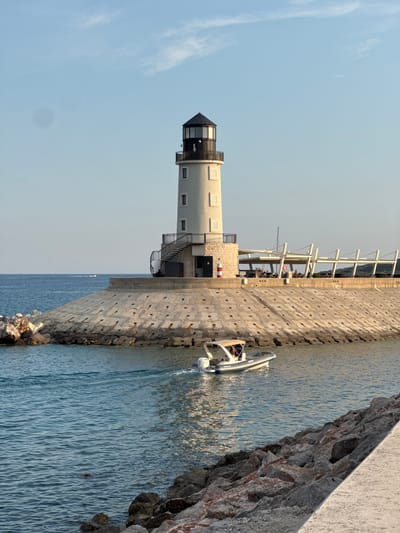 The image shows a lighthouse on a concrete pier in the city of Radovići, Montenegro. A rigid inflatable boat is moving in the water in front of the pier. The lighthouse is white with a black top and has a spiral staircase leading to an upper level. There are people sitting at tables under a covered area behind the lighthouse. The water is calm and blue, and the sky is clear with a few wispy clouds. The pier is made of large concrete blocks and is lined with rocks. The lighting suggests it is late afternoon or early evening.