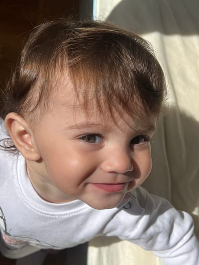 A close-up, tilted, high-angle shot captures a young child with light skin, brown hair, and grey eyes. The child is wearing a white, long-sleeved shirt. The background consists of a cream-colored surface with shadows suggesting a window. The scene does not show anything to suggest the location is Elena, Bulgaria.
