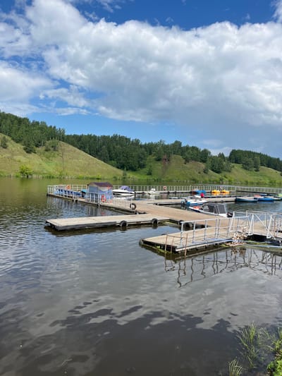 This image depicts a serene lakeside scene at a boat rental station located on the Iset River in Kamensk-Uralsky, Russia. In the foreground, a wooden pier extends into the dark, reflective water, with various small boats and paddle boats docked along its length. The pier is equipped with railings and a small covered structure. In the background, lush green hills covered in trees rise from the opposite bank of the river. The sky above is a mix of blue and white clouds, suggesting fair weather. The overall impression is one of a peaceful recreational area on a summer day.