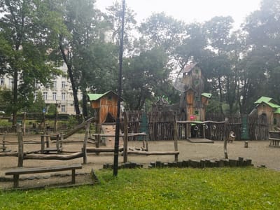The image depicts an outdoor adventure playground in Lviv, Ukraine. The playground features a whimsical, rustic design with wooden structures resembling treehouses and forts, crafted from rough-hewn logs and planks. Green roofs top some of the wooden buildings, adding a splash of color. A wooden slide, climbing walls, and various balance beams and obstacle course elements are visible, suggesting an environment designed for active play and exploration. The ground is covered with gravel and dirt, typical of playgrounds.
In the background, a large, old building with many windows and a light-colored facade can be seen peeking through a canopy of lush green trees. The trees themselves are dense and mature, creating a natural, shaded environment. The sky appears overcast, contributing to a somewhat muted and diffused light, suggesting it might be a cloudy day or early morning/late afternoon.
While there are no people clearly visible in the main playground area, a small, cartoonish figure, possibly a bear dressed in clothing, stands near the center-right of the image, holding a stick. This figure adds to the imaginative and playful atmosphere of the playground. The overall mood is one of natural beauty and adventurous fun, with a slightly overcast or foggy quality to the air, typical of a humid or damp day. No discernible text is visible beyond a small numerical sticker on a lamppost.