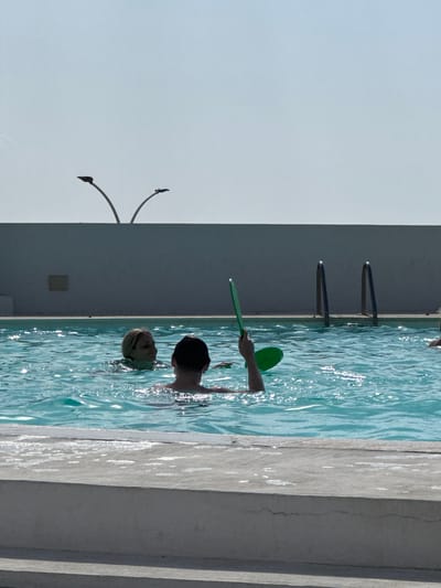 Two children are playing in a swimming pool. One child, with dark hair, is looking away from the camera, their head and shoulders visible above the water. Another child, with blonde hair, is in front of them, also with their head and shoulders above water, looking towards the camera. A green paddle is visible in the water, held by a person whose arm and hand are partially submerged. The pool has a white edge and a concrete walkway next to it. In the background, a dark wall with two metal ladder rungs is visible, partially submerged in the water. The sky is a clear, pale blue, suggesting it is a sunny day. The scene appears to be a private or public swimming pool, possibly at a hotel or residence, given the clean white edge and the presence of ladders. The children appear to be enjoying themselves, with the blonde child's gaze suggesting interaction or a moment of play. No visible text is present in the image.
