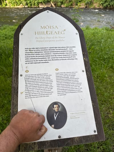 An informational sign in the city of Keila-Joa alevik, Estonia, is depicted. The sign is made of wood and metal and features text in multiple languages, including English, Estonian, and Russian. The text describes the history of the Keila-Joa manor, its previous owners, and its architectural style. A portrait of a man in a suit and tie is shown on the sign, identified as Andrei Shtaken-Schneider, architect of the Keila-Joa manor. In the foreground, a person's hand is visible, obscuring a portion of the sign. The background of the image shows a body of water flowing through a lush green environment with trees and grass.