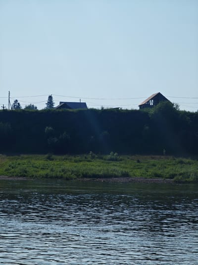 The image shows a wide, tranquil body of water in the foreground, with small ripples reflecting the clear sky. In the middle ground, a densely vegetated embankment rises, covered in lush green grass and various trees. On top of the embankment, silhouetted against the bright sky, are several structures. To the left, a power line with poles is visible, along with what appears to be a house and some trees. Further to the right, another house with a dark roof and a partially constructed, open-framed building with a wooden roof are situated. The overall scene depicts a rural or semi-rural landscape along a waterway on a bright, clear day.