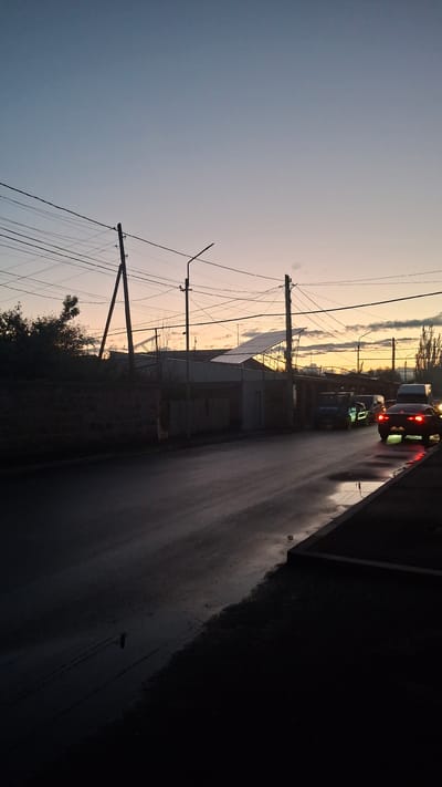 This vertical, eye-level photograph depicts a street scene in Gyumri, Armenia, during twilight. The sky transitions from a pale blue at the top to hues of orange and yellow near the horizon. Overhead, a dense network of power lines is visible, supported by wooden and metal poles.

To the left, a low stone wall runs along the edge of the road, bordered by dark foliage. A paved road, appearing wet with reflections of streetlights and car lights, occupies the lower half of the frame. Several vehicles are present on the road. Closest to the viewer and on the right, a dark-colored car has its brake lights illuminated. Further down the road, a light-colored van and other vehicles are visible, some with headlights on.

Buildings line the right side of the road, including structures with what appear to be solar panels on their roofs. Streetlights are positioned intermittently along the road. The overall impression is that of a residential or light industrial area of Gyumri at the close of day. The lighting suggests a humid or recently rained-on environment.