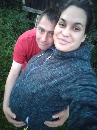 This media file shows a medium shot of a couple posing for a selfie outdoors, likely in a rural setting. The man, wearing a red t-shirt, has his arm around the woman. He is looking at the camera with a slight smile. The woman, in a dark, patterned hooded jacket, is also looking at the camera and smiling. Her expression appears warm and happy. The background consists of lush green foliage and what appears to be a wooden fence and structure, suggesting a garden or yard. The lighting indicates it might be late afternoon or early evening, with soft natural light. The focus is on the couple, with the background slightly blurred, emphasizing their presence and their embrace. The man's hands are around the woman's waist, and her belly is visible, suggesting she is pregnant. The overall mood of the image is intimate and joyful, capturing a moment of connection between the couple. There is no visible text in the image.