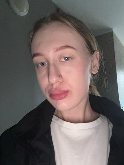 A medium close-up shot of a young woman with fair skin, blue eyes, and light brown hair. She is wearing a white t-shirt and a black jacket. Her face is angled slightly, and she is pouting her lips. A piercing is visible on her nostril. The background is a blurry gray wall. It is impossible to determine the location as Kuleshovka, Russia based on the visual information provided.