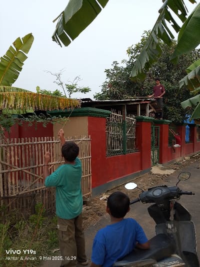 The image shows a scene in Kecamatan Anjatan, Indonesia, located at latitude: -6.417, longitude: 107.978. Three young men are interacting in front of a red brick building with a green gate. The man on the left is holding a string and appears to be attempting to tie it to the gate. He is wearing a green t-shirt and khaki pants. The man on the right is sitting on a black motorcycle, facing the left side of the frame. He is wearing a blue t-shirt. Behind him, a man is climbing on top of a red roof, holding a brown object. He is wearing a red shirt and grey pants. All three men are barefoot. The building is surrounded by a bamboo fence and several trees. The sky is overcast with gray clouds. The image was taken on a sunny day. The ground is dusty and littered with debris. The ground and surrounding area are relatively clear of foliage. The image is taken from a slightly elevated angle, looking down at the scene. The image was taken with a smartphone, as evidenced by the watermark on the bottom right corner of the image. The watermark indicates that the image was taken with a Vivo Y19s smartphone on May 9, 2023, at 4:29 PM.