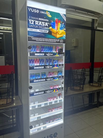 This media file captures a retail display of vaping products, likely inside a convenience store or electronic cigarette shop in Makassar, Indonesia. The primary subjects are the various vaping devices and e-liquid cartridges neatly arranged in a brightly lit, vertical display case.

The scene is set in an indoor retail environment. The display case, illuminated by white LED lights, showcases products from brands such as "VUSE CIRCLE K" and "RELX". On the left side of the display, a large promotional banner highlights "VUSE CIRCLE K" and advertises "3 TAHUN BERSAMA" (3 years together) with "12 RASA UNTUK DICOBA!" (12 flavors to try!). Below this, prices are displayed, such as "KINI DARI Rp54.000" (Now from Rp54.000). The banner also includes a warning: "Produk ini hanya untuk 21 tahun ke atas, mengandung nikotin dan dapat menyebabkan ketergantungan. Dilarang menjual dan memberi kepada orang di bawah usia 21 tahun dan perempuan hamil." (This product is only for 21 years and above, contains nicotine and can cause addiction. It is forbidden to sell or give to people under 21 years of age and pregnant women.).

Various "VUSE GO" models are prominently featured in the display, including "VUSE GO 700", "VUSE GO 1000", "VUSE GO RELOAD", and "VUSE GO 3000", each with accompanying prices like "Rp62.000", "Rp76.500", "Rp54.000", and "RP162.000" respectively. Other items, some appearing to be RELX products, are also visible. There are no people clearly visible in the image, suggesting the photograph was taken when the store was not busy or the photographer was focusing solely on the products. The lighting indicates it could be daytime or that the interior lighting is quite strong. The background shows glimpses of store shelves and structures, possibly including metal racks. The overall impression is that of a commercial display designed to attract customers and inform them about available vaping products and their prices.