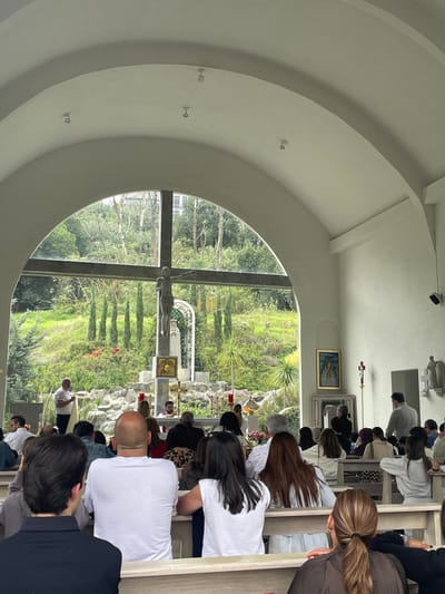 The image depicts the interior of a chapel in Huixquilucan de Degollado, Mexico. Several rows of people are kneeling or sitting in pews facing forward. A large, arched window reveals an outdoor religious scene with a prominent crucifix and a statue, surrounded by greenery and foliage. Inside the chapel, religious artwork hangs on the wall. The ceiling features a series of concentric arches.