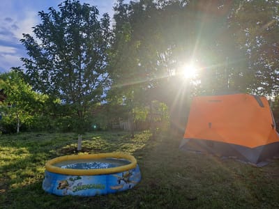 This media file depicts a sunny outdoor scene in a rural or suburban setting, identified as Полянки, Russia. The primary subjects are an orange and gray camping tent and a blue and yellow inflatable kiddie pool.
The tent, branded "TopCamping", is positioned on the right side of the frame and features a metal chimney-like pipe extending upwards from its top. To the left, the kiddie pool is partially filled with water and rests on the grass. It is decorated with cartoon airplane characters and the text "El Chupacabra" and "Disney Planes".
The scene is set in a grassy area, likely a backyard or garden, surrounded by several trees with lush green foliage. The ground exhibits varying lengths of grass, suggesting a natural and perhaps recently maintained space. Bright sunlight streams into the scene from the right, creating a prominent lens flare and casting long shadows across the ground, indicating either early morning or late afternoon. The sky, visible in parts through the tree canopy, is a clear blue with some scattered clouds. In the background, sections of a wooden fence and a wire fence are discernible. No people are present, but the setup of the tent and pool suggests activities related to leisure, camping, or children's play. The overall mood is peaceful and bright.