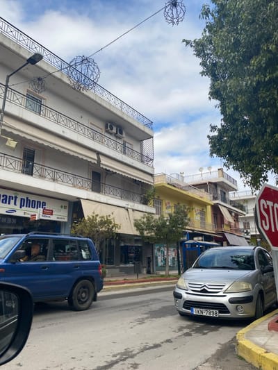 This image captures a tilted street scene in Koridallos, Greece, under a partly cloudy sky with patches of blue. The photograph appears to be taken from inside a vehicle, with a side mirror visible in the bottom right.
On the right side of the street, a silver Citroën C3 is parked with its front wheels partially on a yellow curb. Its Greek license plate, "IKN-7898," is clearly visible. A red octagonal "STOP" sign is positioned above this car. Further down the street, a blue SUV, possibly a Suzuki Jimny given the rear-mounted spare tire, is seen.
Buildings line the left side of the street, featuring multiple stories with balconies adorned with dark metal railings. Several air conditioning units are mounted on the facades of these predominantly light-colored buildings. Awnings extend over the ground-floor shops. One shop's sign clearly advertises "Smart phone repair" and includes logos for brands such as Samsung, Microsoft, Nokia, and Sony. Further along, another building with a vibrant yellow facade is visible.
Above the street, several decorative spherical lights are strung across, potentially for street beautification or festive purposes. Green tree foliage is prominent in the upper left corner and intermittently along the street. The scene depicts a typical daytime urban environment, bustling with parked vehicles and street decorations.