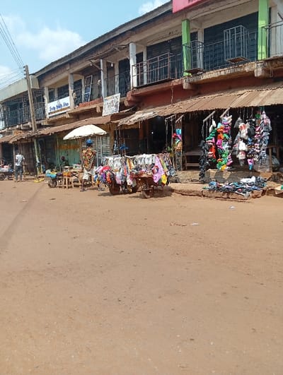 The image displays a street-level scene of a commercial area in Nsukka, Nigeria, under clear daylight. The foreground and midground consist of an unpaved, reddish-brown dirt road with scattered small stones.
In the background, a continuous row of two-story buildings lines the street. These structures feature ground-level shopfronts, many equipped with weathered, corrugated metal awnings, some exhibiting rust. The upper stories display balconies with dark metal railings. One section of the upper floor on the right features light green painted pillars. Visible signage includes "Sweetly Healthy Shop" on an upper floor and another less legible sign on a lower level.
Three individuals are discernible. On the far left, a person in a white t-shirt stands near a small cart. Centrally, a person in a brightly patterned shirt and dark headwear stands beneath a white parasol, attending to a merchandise cart displaying various small items. A third person is partially visible within a shop entrance further left.
Commercial displays are prominent at street level. On the right, a stall features numerous pairs of multicolored sandals and slippers hanging from suspended racks, with additional footwear arranged on the ground below. Another cart, laden with items, is positioned centrally near the parasol-shaded vendor. Electrical utility poles and overhead wires are visible in the upper left of the frame. The overall scene depicts a bustling, informal market environment.