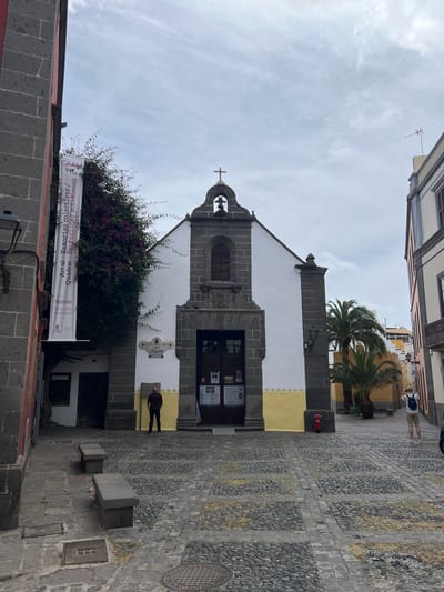 Here is a detailed description of the image:
The image depicts a street scene in the city of Las Palmas De Gran Canaria, Spain, featuring a church situated between buildings. The architecture of the church is characterized by a white facade with dark stone detailing. It has a bell tower surmounted by a cross. A banner hangs on the left side of the church, partly obscured by foliage, with text reading "Art al Memorias colectivas" and exhibition dates "19.03.2026 - 30.08.2026." A small plaque with the word "COLOR" is mounted on the church wall.
The street is paved with patterned stones. A red fire hydrant is visible on the right side of the church. A few people are present: one is walking near the church entrance, and others are further up the street. The buildings on either side of the church show a mix of architectural styles and colors. Palm trees are also visible.
The sky appears overcast. The scene is typical of an urban environment with a mix of historical architecture and modern elements.