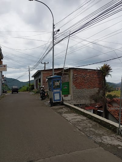 The image shows a street in Kecamatan Kutawaringin, Indonesia, at the coordinates -6.987 latitude, 107.531 longitude. The scene features a paved road with a small food cart parked on the right-hand side. The cart is painted blue and has a white sign with text that is not visible. A motorcycle is parked in front of the cart. Behind the cart is a brick wall. The wall is made of red bricks and painted white. Behind the wall, there is a small green roof and a patch of green grass. Above the street is a cloudy sky with a streetlight extending from the left side of the frame and a  large number of electrical wires that crisscross the scene. The image was captured from the point of view of a person standing on the side of the road looking down the street. There are no people in the image.  The street is mostly empty, with only a single dark-colored car visible in the distance.
