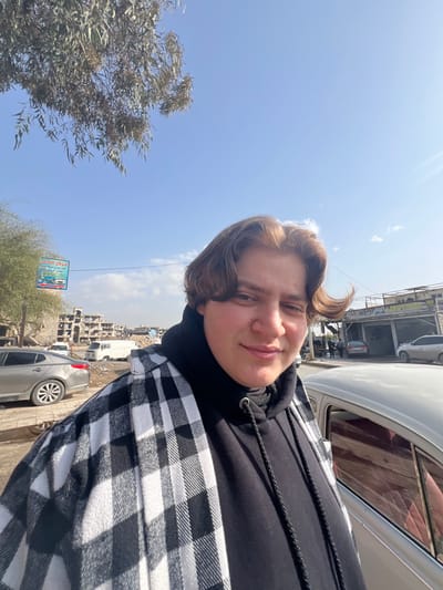 A young individual with light brown hair, wearing a black hooded sweatshirt beneath an open black and white plaid shirt, stands in the foreground, facing the camera with a slight smile. A light-colored vehicle, possibly a Volkswagen Beetle, is partially visible to their right. The scene is located in Joubar Municipality, Syria. The background reveals a damaged urban environment featuring multiple multi-story buildings in various states of disrepair, with visible structural damage, exposed rebar, and broken windows. Several vehicles, including a gray sedan and a white van, are parked on a street or shoulder composed of dirt and pavement. To the left, a blue and white billboard displaying Arabic text, including "مركز العالم" (World Center), is visible. Overhead, the sky is clear and blue with scattered white clouds, and the leaves of a tree are visible in the upper left. The ground in the midground and background appears to be a mix of unpaved areas, debris, and functional road surfaces, indicating an area with significant prior destruction and ongoing activity.