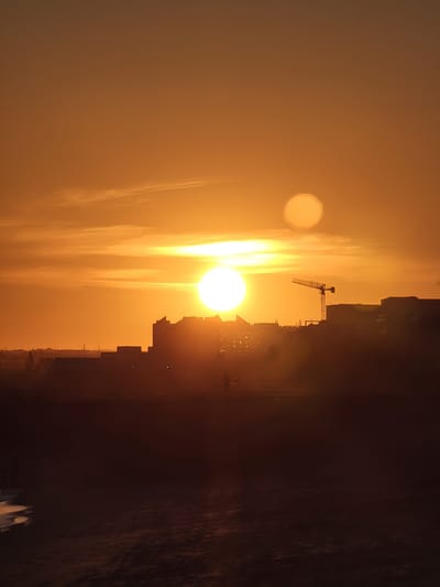 The image captures a dramatic sunset over the city of Quarteira, Portugal. The sky is ablaze with shades of orange and yellow, with two distinct suns visible. The larger sun, partially obscured by clouds, casts a warm glow, while a smaller, fainter sun hovers above it. In the foreground, a silhouette of the city skyline is visible, dominated by buildings and a construction crane. The lower part of the image shows a dark, sandy beach meeting the gentle waves of the ocean. The overall scene is one of natural beauty and urban development, bathed in the warm, fading light of the setting sun.