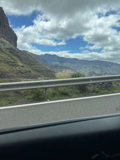 The photograph, taken from inside a vehicle, shows a roadside scene in Mogán, Spain. A grey guardrail separates the road from a steep, green, mountainous terrain. The road is asphalt, marked with a white line along the edge. In the distance, layered mountain ridges are visible, ascending into a sky partially obscured by white, puffy clouds with patches of blue sky showing through. The view suggests a high altitude or elevated road position. The vehicle frame is visible, partially blocking the right portion of the photograph. The composition emphasizes the contrast between the man-made road and the natural, rugged landscape.