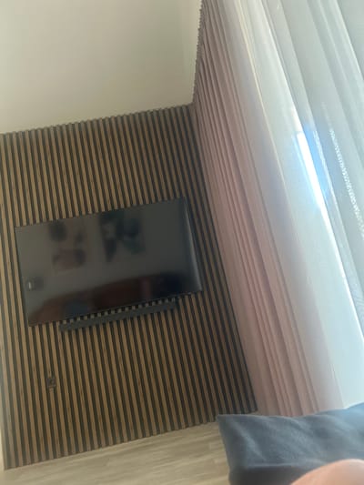 The image depicts an indoor space, specifically identified as being in Markovo, Bulgaria. The primary focus is a wall adorned with vertical, light-brown wood slatted paneling, featuring darker grooves between the slats. A large, black flat-screen television is mounted centrally on this slatted wall, with a matching black soundbar positioned directly beneath it. The television screen is off and reflects blurred, indistinct shapes, appearing to be two human figures or faces positioned towards the upper left and right quadrants of the screen. Below the TV and soundbar, a dark electrical outlet is visible on the slatted wall in the lower-left quadrant.

To the right, the slatted wall transitions into an interior corner, revealing a window covered by a double layer of curtains. The inner layer consists of a pleated, light pink or mauve fabric, while the outer layer is a sheer, translucent white material. Bright natural light, indicative of daylight, streams intensely through the sheer curtain, creating a vertical band of luminosity.

The ceiling, visible at the top of the image, is plain and light-colored. The floor, visible in the lower-left and central portions, appears to be a light-toned wood or laminate. In the immediate foreground, a grey, soft textile object, likely a pillow or blanket, occupies the bottom-right corner, partially obscuring the view of the floor, and a small segment of human skin is visible at the extreme bottom right. No explicit human actions or interactions are discernible beyond the reflections in the television.