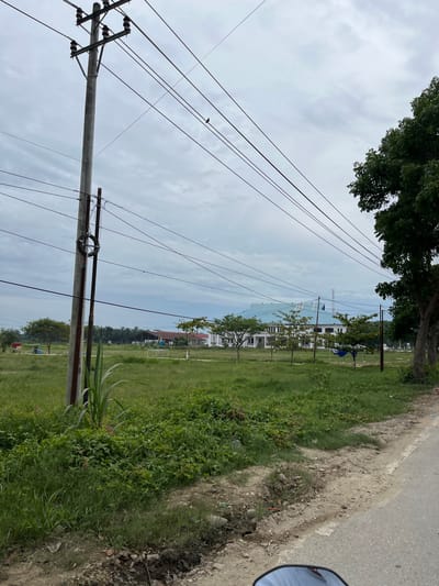The image depicts an outdoor scene in Bundar, Indonesia, with a focus on a utility pole and its overhead wires. The pole, made of concrete, stands to the left of the frame and supports multiple electrical wires. A coil of black cable is attached to the pole. In the background, a white building with a teal-colored roof is visible, surrounded by green grass and scattered trees. The sky is overcast with clouds. A paved road runs along the right side of the image, and the edge of what appears to be a motorcycle's rearview mirror is visible in the lower right corner. The ground in front of the building is grassy, with some patches of dirt and debris.