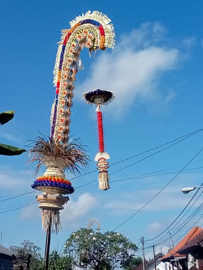 The image depicts two elaborately decorated, tall, bamboo-like structures against a clear blue sky with scattered white clouds. These structures appear to be ceremonial decorations, possibly for a Balinese festival. The left structure is taller and curves upwards, adorned with colorful floral arrangements and intricate woven patterns in red, blue, yellow, and white. It is topped with a large, fan-shaped ornament. Below this, it features a woven basket-like structure with layered, colorful floral elements and a plume of dried grass.
To the right, a shorter, hanging decoration is suspended from a pole. This decoration has a dark, circular top, followed by a long, red, textured cylindrical section, and a smaller, elaborately woven base with straw-like tassels. The background includes a few green leaves from trees on the left, some power lines crisscrossing the sky, a streetlamp on the right, and rooftops of buildings in the lower right corner, suggesting an outdoor, possibly urban or semi-urban, environment. The presence of these structures and the overall aesthetic strongly suggest a location in Bali, Indonesia, likely Blahbatuh, known for its cultural celebrations and art. There are no visible people in the image. The scene captures a moment of festive decoration, highlighting the craftsmanship and vibrant culture of the region.