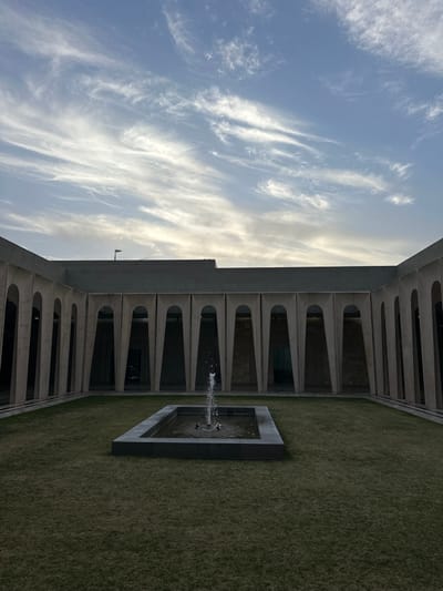 The scene depicts an unoccupied, manicured inner courtyard of a modern institutional facility located within the Galil Elyon Regional Council, Israel. The courtyard is enclosed by a building featuring a continuous colonnade composed of tall, repetitive, light-toned, vertically elongated arches with a trapezoidal-like form. Above the arches, the building's upper wall section is flat and dark gray. The ground within the courtyard is covered by well-maintained green grass. Centrally positioned on the grass is a rectangular, dark gray fountain basin from which a single jet of water actively projects upwards. The sky above is light blue, characterized by scattered, wispy white cirrus clouds. The environment is illuminated by soft, natural light, suggesting late afternoon or early evening conditions. No individuals, animals, or additional objects are visible in the frame.