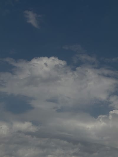 The image is a photograph of a sky filled with clouds, with a deep blue sky visible above. There are varying densities of clouds, ranging from wispy, white formations to larger, more substantial gray clouds that appear to be closer to the viewer. The lighting suggests it is daytime, with sunlight filtering through and illuminating parts of the clouds. There are no visible buildings, landmarks, or geographical features that would place this image specifically in Chirchik, Uzbekistan. The photograph solely depicts a natural sky scene.