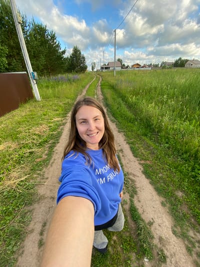 A woman is taking a selfie on a dirt road in a rural area, likely the village of Garevaya, Russia. She is smiling and wearing a blue sweatshirt with white lettering that reads "My mom I'm fabulous". The road is flanked by tall green grass and wildflowers. In the background, there are trees, houses, and power lines under a cloudy blue sky.