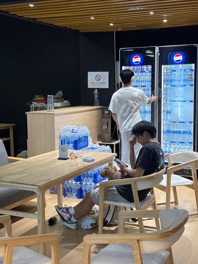 Here is a description of the image:
**Location:** The image appears to be taken indoors at a business or restaurant in the Nan'an District, China.
**Scene:** The scene is a casual setting, likely a break room or refreshment area.
**People:** There are two people in the image, both young men of East Asian descent.
* One is wearing a white shirt and stands at a Pepsi vending machine.
* The other is sitting in a wooden chair, wearing a black shirt and gray shorts. He is using a smartphone.
**Objects:**
* Pepsi vending machines: There are two large Pepsi vending machines stocked with bottled water.
* Wooden chairs and tables: There are several wooden chairs and a long wooden table.
* Bottled water: Numerous bottles of water are piled on the table.
* Smartphones
* Dog: A small, brown, poodle-type dog is seen by the chair.
**Actions:**
* One man is interacting with the Pepsi vending machine.
* The other man is sitting down and using his smartphone.
**Interactions:** The two men seem to be together, possibly taking a break. The second man has his attention on his phone while the first man interacts with the vending machine.
**Environment:** The setting is clean and well-lit. The walls are a combination of black and wood paneling.