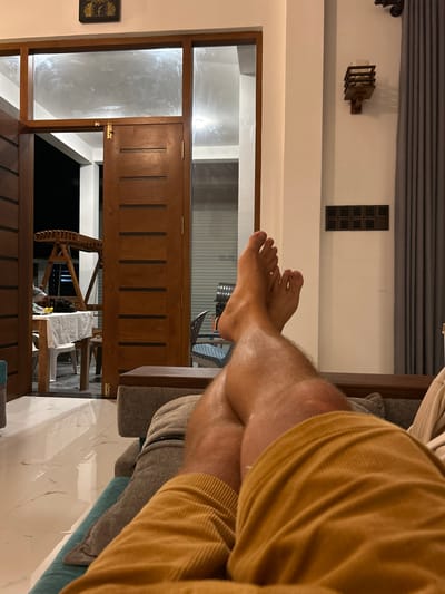 The image shows a person's legs and feet resting on a sofa, with their body out of frame. The person is wearing an orange ribbed sweater and what appears to be shorts. The setting is indoors, likely a living room, with large wooden doors and glass windows dominating the background. Through the glass, a glimpse of an outdoor area can be seen, possibly a patio or garden, with some furniture and structures suggesting a resort or a modern home. The lighting is dim, indicating it's either evening or nighttime. The overall ambiance is relaxed and comfortable, with the person in a reclined position. There are decorative elements on the wall, including a modern wall sconce and a patterned panel. A clock is visible on the left side of the frame. The floor appears to be tiled with a marble-like pattern. The orientation of the image is vertical, with the scene appearing sideways.
