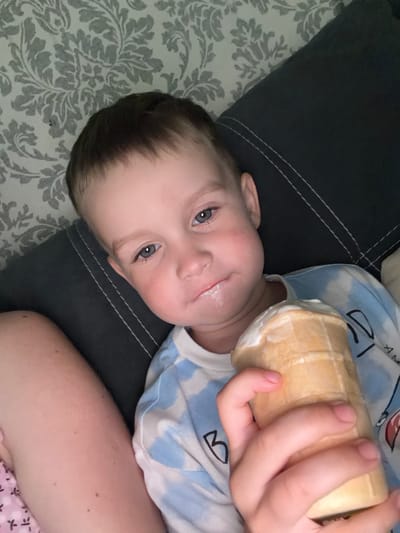 The media file shows a close-up, rotated view of a young child lying on a surface patterned with grey floral motifs and dark grey fabric. The child, with fair skin, blue eyes, and dark hair, is looking upwards. There is a slight sheen of moisture around their mouth, suggesting they have recently eaten something, possibly ice cream, as a partially eaten ice cream cone is held in their right hand. The child is wearing a light blue and white tie-dye patterned shirt with black line drawings of clouds and letters. The setting appears to be indoors, likely a comfortable domestic environment, given the pillows or cushions and the relaxed posture of the child. The lighting suggests it could be daytime or evening, with no strong indications of natural light. The overall mood is one of innocent enjoyment, with the child seemingly content and engaged with the ice cream cone. No visible text is present on the child or the immediate surroundings, other than the stylized drawings on the shirt. The provided location context of Bila Tserkva, Ukraine, places this scene in that specific geographical region.