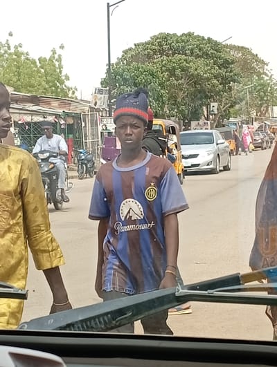 The image depicts a busy daytime street scene in Hotoro North, Nigeria, observed from inside a vehicle, indicated by the dashboard and windshield wiper in the lower frame.
In the immediate foreground, to the left, is the partial profile of an individual wearing a gold-colored, patterned traditional garment and a thin, light-colored bracelet on the left wrist. Centrally positioned is a young male, mid-shot, looking directly forward. He wears a blue and maroon striped t-shirt with a white 'Paramount' logo and mountain graphic, and a yellow circular emblem with black detailing on the left sleeve. He also wears a dark blue knitted cap with red stripes and a pom-pom.
The street beyond features active pedestrian and vehicular traffic. A male subject riding a black motorcycle, dressed in a light blue traditional tunic, pants, dark sunglasses, and a dark cap, proceeds from left to right. Behind him, a silver sedan (Toyota Corolla) and two yellow motorized rickshaws are visible in traffic. Further down the road, multiple pedestrians are present, including one in a pink and white patterned dress.
To the left, several commercial structures with corrugated metal roofs and green barred windows display goods. Visible signs include "HOTEL" and "AL HMEED S.S.". To the right, another commercial building has a partially visible sign reading "2 SISTER M". Large, leafy green trees dominate the upper half of the background, along with a black streetlight pole. A poster featuring a male portrait is affixed to a pole or tree in the midground right. The sky is bright and hazy, indicating daylight conditions.